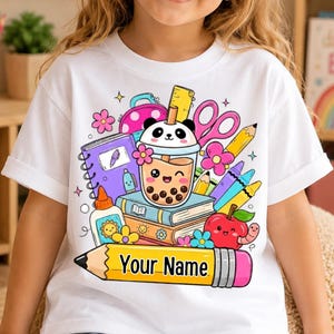 May include: A white t-shirt featuring a colourful graphic of school supplies including a boba tea cup with a panda face, books, scissors, glue, and an apple with a worm. A large yellow pencil at the bottom contains the text Your Name.