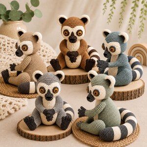 May include: Five crocheted lemur plush toys in various colors including brown, tan, blue, gray, and sage green. Each lemur features a cream-colored face, dark brown nose, amber-colored eyes, and a long, striped tail, all sitting on wooden discs.