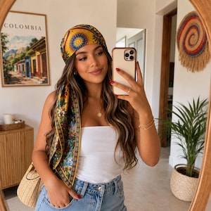 May include: A square silk headscarf featuring a vibrant yellow, blue, and red pattern with sun motifs and floral designs. The scarf is tied around the head with one corner draped over the shoulder, showcasing a detailed border and intricate illustrations.