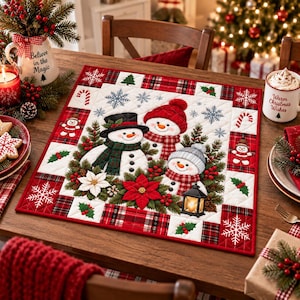 May include: A square quilted placemat featuring three snowmen, red poinsettias, and green pine branches. The border displays a red and black plaid pattern with white snowflakes, candy canes, and holly leaves. The design includes a black lantern.