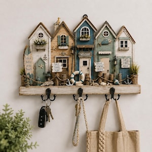 May include: A wall-mounted key holder featuring a row of five miniature beach-themed houses in white, tan, and blue. The houses include signs reading Life is Better at the Beach, Salt Sand Surf, Welcome, Relax Unwind Enjoy, and Good Vibes Only, with three black hooks.