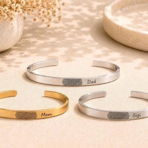 Könnte beinhalten: Drei offene Armreifen, zwei in silberfarbenem und einer in goldfarbenem Metall. Jeder Armreif zeigt ein lasergraviertes Fingerabdruck-Design, gefolgt von einem Namen: Dad auf einem silbernen Armreif, Mom auf dem goldenen und Gigi auf dem anderen.