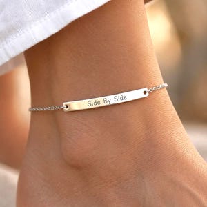 May include: A silver-toned metal anklet featuring a thin, delicate chain and a central horizontal rectangular bar. The bar is engraved with the black text Side By Side in a simple, clean font. The piece is designed to wrap around the ankle.
