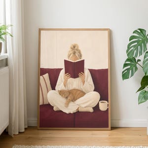Puede incluir: Una lámina de arte enmarcada que muestra a una persona con el pelo rubio recogido en un moño, vestida con ropa de estar color crema, sentada con las piernas cruzadas en un sofá rojo oscuro, leyendo un libro rojo oscuro con un gato atigrado naranja.
