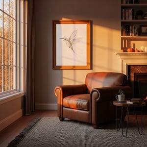 May include: A brown leather armchair with rolled arms, positioned next to a small round wooden side table holding a white mug. Above the chair hangs a framed wall art piece featuring a delicate line drawing of a hummingbird in flight.
