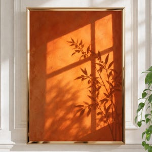 May include: A rectangular wall art piece in a thin gold frame featuring a textured, vibrant orange background. The artwork displays a dark, sharp shadow cast by a window frame and a leafy plant branch, creating a high-contrast silhouette effect.