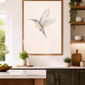 May include: A framed wall art print featuring a delicate, minimalist line drawing of a hummingbird in flight. The artwork uses fine black ink strokes on a light cream background, capturing the bird's wings and long tail feathers in motion.
