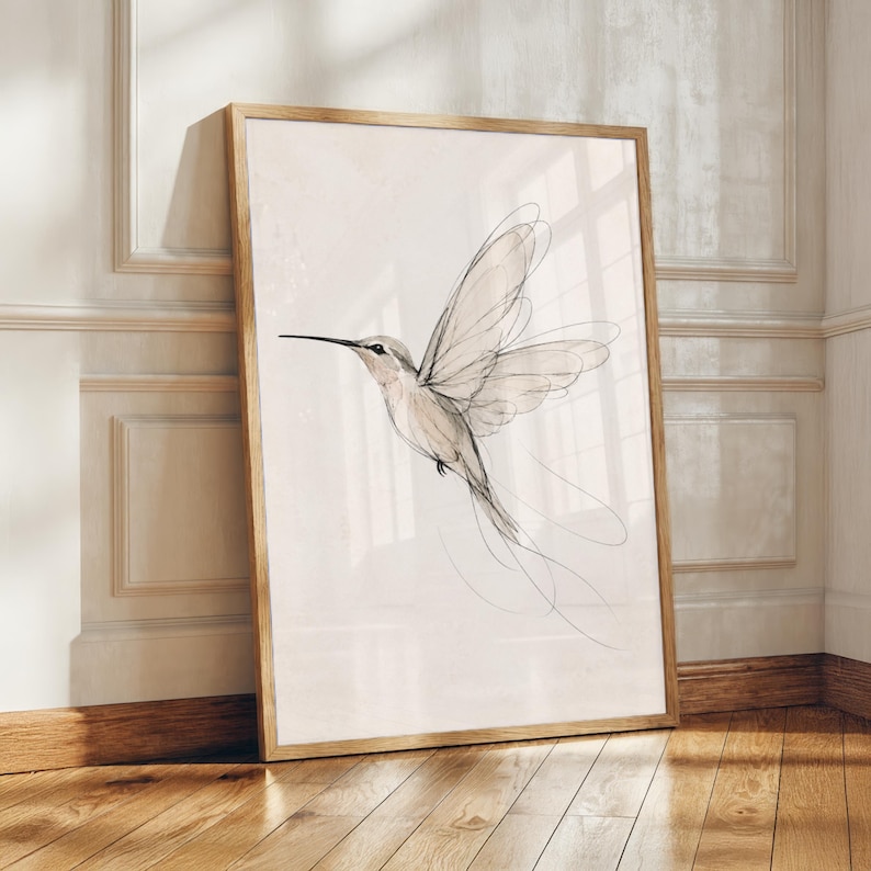May include: A framed wall art print featuring a delicate, minimalist line drawing of a hummingbird in flight. The artwork uses fine black ink strokes against a soft, pale beige background, capturing the bird's wings and long tail feathers.