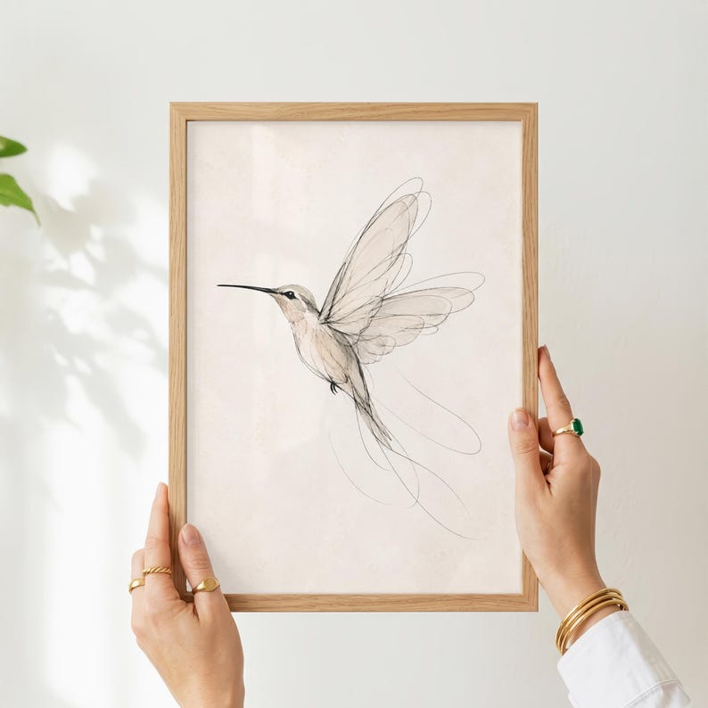 May include: A framed wall art print featuring a delicate, minimalist line drawing of a hummingbird in flight. The artwork uses fine black ink strokes on a light, off-white background, captured within a simple, light-toned wooden frame.