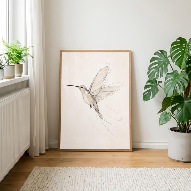 May include: A framed wall art print featuring a delicate, fine-line sketch of a hummingbird in flight. The drawing uses soft, thin black lines against a light beige background, capturing the bird's wings and long tail feathers in motion.