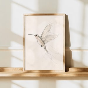 May include: A framed wall art print featuring a delicate, fine-line sketch of a hummingbird in flight. The bird is rendered in soft charcoal and light brown tones against a pale, cream-colored background, encased in a light wood frame.