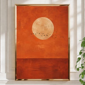 Pode incluir: Uma impressão de arte de parede emoldurada com uma grande lua circular pálida centrada contra um céu laranja profundo e texturizado. Um bando de pequenos pássaros pretos é silhuetado em voo sobre a parte inferior da lua. O terço inferior da impressão retrata uma linha de horizonte laranja escura e sólida, representando um corpo de água calmo.