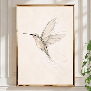 May include: A framed wall art print featuring a delicate line drawing of a hummingbird in flight. The bird is rendered in fine black ink strokes against a solid, light beige background, captured within a thin, metallic gold-colored frame.