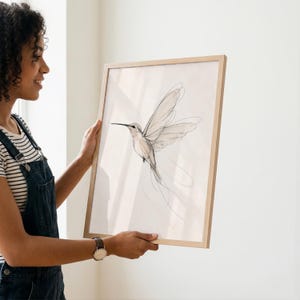 May include: A framed wall art print featuring a delicate, minimalist line drawing of a hummingbird in flight. The artwork uses thin, fluid black ink strokes on a light, off-white background, encased in a simple, light-toned wooden frame.