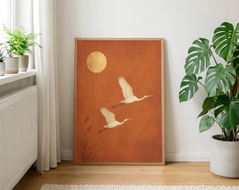 May include: A rectangular wall art print in a light wood frame featuring a burnt orange background with two white cranes in flight, a golden circular sun, and thin black reeds at the bottom left corner, displayed in a bright interior space.