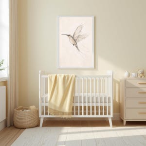 May include: A framed wall art print featuring a delicate black line drawing of a hummingbird in flight against a light cream background, displayed above a white wooden crib with a soft yellow throw blanket draped over the side rail.