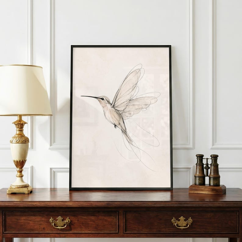 May include: A framed wall art print featuring a delicate, minimalist line drawing of a hummingbird in flight. The bird is rendered in fine black ink strokes against a soft, cream-colored background, encased in a simple black rectangular frame.