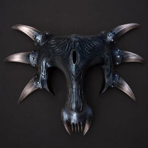 May include: A dark, metallic-finished mask featuring a symmetrical, organic shape with six prominent, curved horn-like protrusions extending from the sides. The central section includes a vertical eye-shaped opening and a lower jaw with sharp teeth.