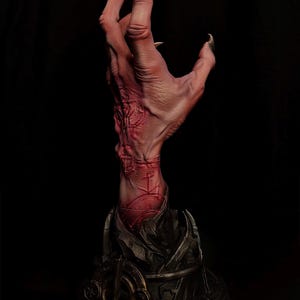 May include: A sculpted hand reaching upward from a dark, circular base. The hand features pale, textured skin with prominent red, etched symbols and long, sharp, dark-tipped fingernails. The base is metallic gray with intricate, carved details.