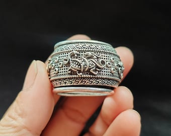 May include: A silver-colored, cylindrical ring with intricate embossed designs. The ring features detailed patterns and a series of small, circular cutouts. The design includes a repeating geometric pattern and a central motif of a stylized creature.