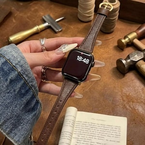 May include: A brown leather watch band with a gold buckle is attached to a black smartwatch. The watch displays the time 18:48. The watch band is on a brown surface.