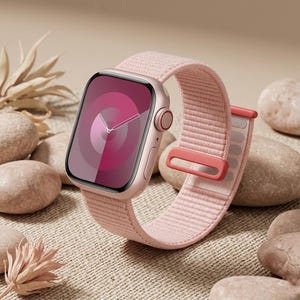 Peut inclure: Une montre connectée rose clair avec un écran rectangulaire et un bracelet en tissu tissé assorti. La montre présente un cadran numérique avec des motifs circulaires concentriques et un bouton couronne latéral. Le boîtier a une finition rose métallique.