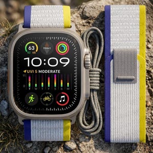 Op de afbeelding: Een titaniumkleurige smartwatch met een digitaal display dat 10:09, UVI 5 en diverse activiteitspictogrammen toont. Het horloge heeft een geweven nylon band met witte, paarse en gele strepen, vergezeld van een losse bijpassende band en een grijs koord.