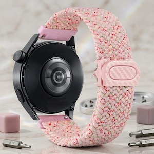 Puede incluir: Un reloj inteligente negro con una carcasa trasera circular y una correa de tela trenzada rosa. La correa presenta un sutil estampado floral y una hebilla de ajuste de plástico rosa claro con una textura de fibra de carbono.