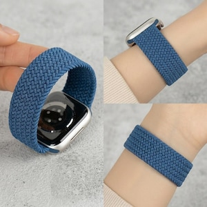 May include: A blue braided elastic watch band featuring a woven texture. The band is shown attached to a silver-toned smartwatch case with a black circular sensor array on the back, and also displayed wrapped around a wrist for a secure fit.