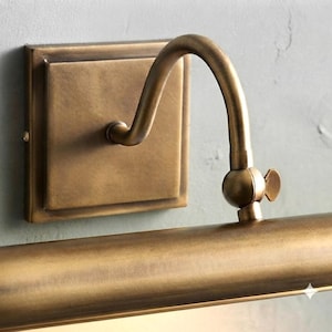 May include: A wall-mounted picture light with an antique brass finish. The fixture features a square backplate, a curved arm, and a cylindrical light housing with an adjustable swivel joint secured by a small wing nut screw on the side.