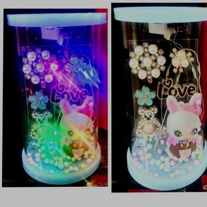 May include: A cylindrical clear display case containing a small rabbit figurine, a teddy bear charm, a flower-shaped ornament, and a decorative piece with the word Love. The interior is filled with small white spheres and illuminated by LED lights.