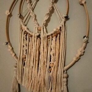 Può includere: Una decorazione da parete composta da due cerchi in legno intrecciati con corda in macramè color bianco sporco. Il design presenta motivi a nodi, perline in legno naturale e piccoli accenti floreali chiari fissati ai cerchi, con lunghe frange.