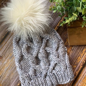 May include: A gray knitted beanie with a white faux fur pom pom. The beanie has a cable knit pattern.
