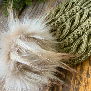 Könnte beinhalten: Eine grüne Strickmütze mit einem flauschigen weißen Bommel. Die Mütze hat ein Zopfmuster.