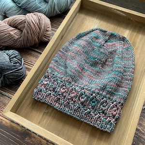 May include: A knitted hat with a teal, pink, and gray color pattern. The hat has a folded brim with a textured stitch.