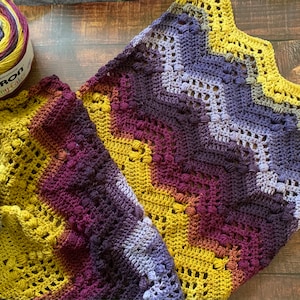 May include: A crocheted blanket with a chevron pattern in shades of purple, yellow, and brown. The blanket is made with Caron Skinny Cake yarn.