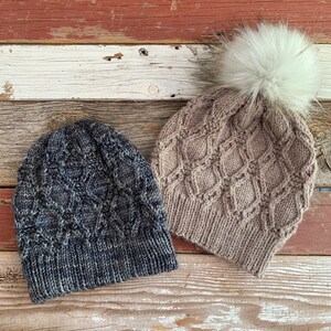 May include: Two knitted beanies. One is a dark blue with a diamond pattern, and the other is tan with a diamond pattern and a white faux fur pom-pom on top. The beanies are on a weathered wood surface.