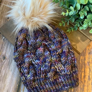 May include: A knitted beanie with a cable knit pattern in shades of purple, brown, and blue. The beanie has a fluffy faux fur pom pom on top.
