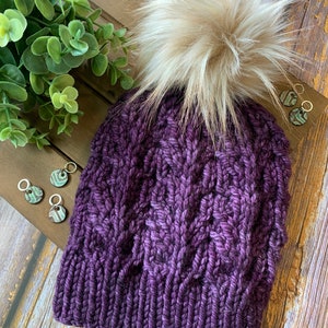 May include: A dark purple knitted hat with a cable knit pattern and a large, fluffy, cream-coloured pom-pom. The hat is displayed on a wooden surface, with small decorative earrings nearby.