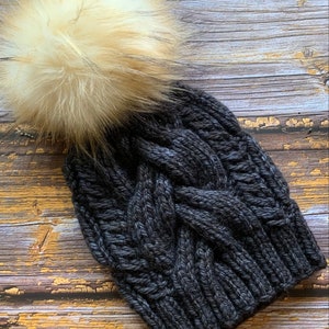 May include: A dark gray knitted beanie with a large, fluffy, cream-colored pom pom.