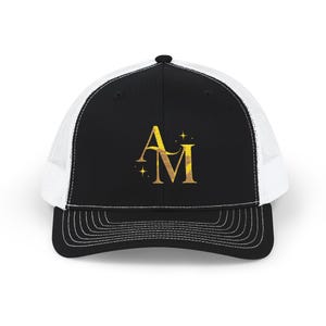 May include: Black and white trucker cap featuring a gold-coloured "AM" monogram and star accents. The hat has a black front panel, a white mesh back, and a curved peak with white stitching. A stylish accessory.