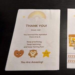 Op de afbeelding: Een wit papieren certificaat met een pastelkleurige regenboog en wolken bovenaan. De tekst luidt: THANK YOU! Great Job! You learned the alphabet from A to Z. Keep practicing, keep learning, and keep having fun! Bevat een hartje, een uil en een ster.