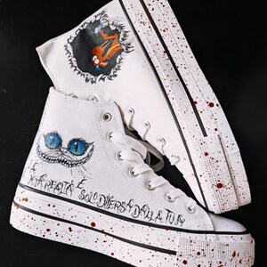 Könnte beinhalten: Weiße High-Top-Plateau-Sneaker mit roten Farbspritzern auf Sohlen und Obermaterial. Ein Schuh zeigt eine handgemalte orangefarbene Kreatur in einem dunklen Loch, der andere ein blauäugiges Katzengesicht und italienischen Text in schwarzer Tinte.