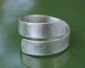 May include: A silver-colored wrap ring with a textured surface. The ring has a modern, minimalist design, with a band that wraps around the finger. The ring is shown on a green surface.