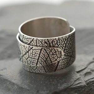 Könnte beinhalten: Ein silberner Ring mit einem Blattdesign. Der Ring besteht aus zwei miteinander verbundenen Bändern. Das Blattdesign ist detailliert und filigran.