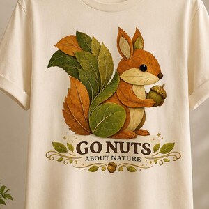 Puede incluir: Una camiseta de manga corta color crema con un gráfico de una ardilla compuesta por hojas de otoño, sosteniendo una bellota. Debajo de la ardilla, el texto dice GO NUTS ABOUT NATURE, enmarcado por hojas decorativas y una pequeña bellota.