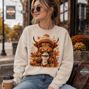 Può includere: Una felpa girocollo color crema con la grafica di una soffice mucca Highland che indossa un berretto di lana e una sciarpa, mentre tiene in mano una tazza con la scritta Pumpkin Spice Latte, circondata da foglie autunnali e una zucca.