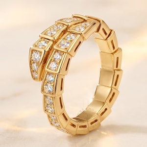 May include: A gold-toned ring shaped like a coiled snake, featuring a segmented body design. The head and upper portion of the snake are encrusted with numerous clear, sparkling round-cut stones, while the rest of the band remains smooth gold.