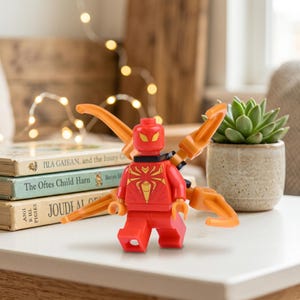 Peut inclure: Une figurine Lego rouge avec un emblème d'araignée doré et des bras mécaniques orange. Elle est posée sur une surface blanche près de livres et d'une plante succulente. Les livres sont intitulés "IIIA GAIEAN" et "The Oftes Child Harn".