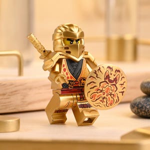 Op de afbeelding: Een gouden ninja-actiefiguur met een zwaard en schild. De figuur draagt een gouden helm, harnas en een masker. Het schild heeft een rood en oranje ontwerp. De figuur staat op een lichtbruin oppervlak.
